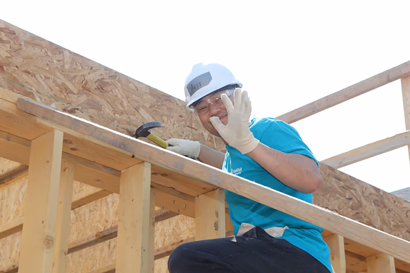 A young man wearing a white hard hat and holding a hammer waves while crouching on an in-progress building