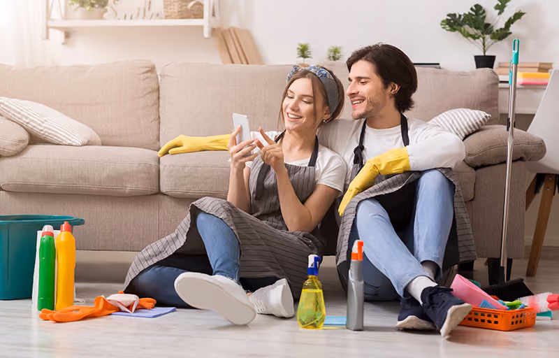 A man and woman lean against a couch while sitting on the floor, surrounding by cleaning supplies, while looking at the woman's phone