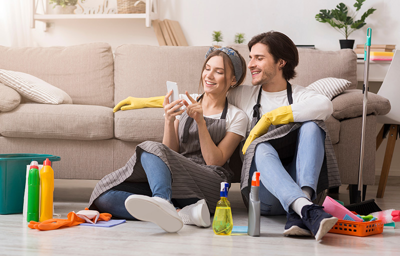 A man and woman lean against a couch while sitting on the floor, surrounding by cleaning supplies, while looking at the woman's phone