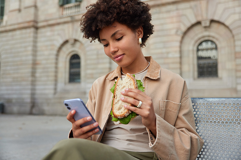 A woman holds a sandwich, looking down at a cellphone in her other hand while seated