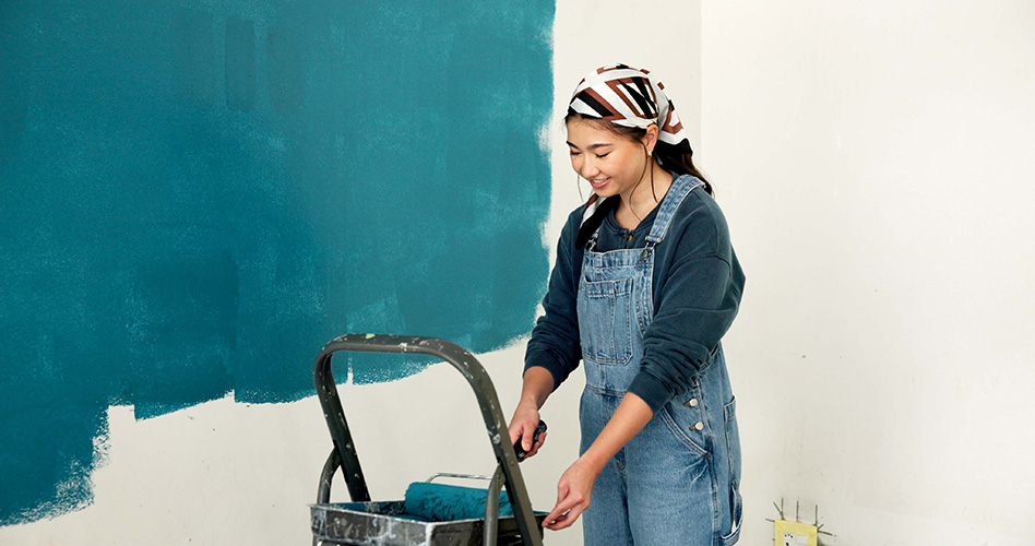 A young woman in overalls and a bandanna looks down at a paint roller she's holding, resting it on a ladder next to a half-painted wall