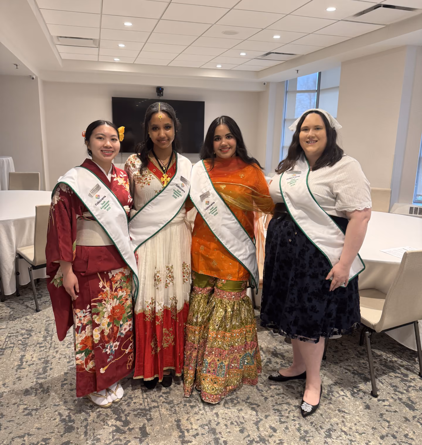 Four women in various cultural attire stand together in a line, smiling