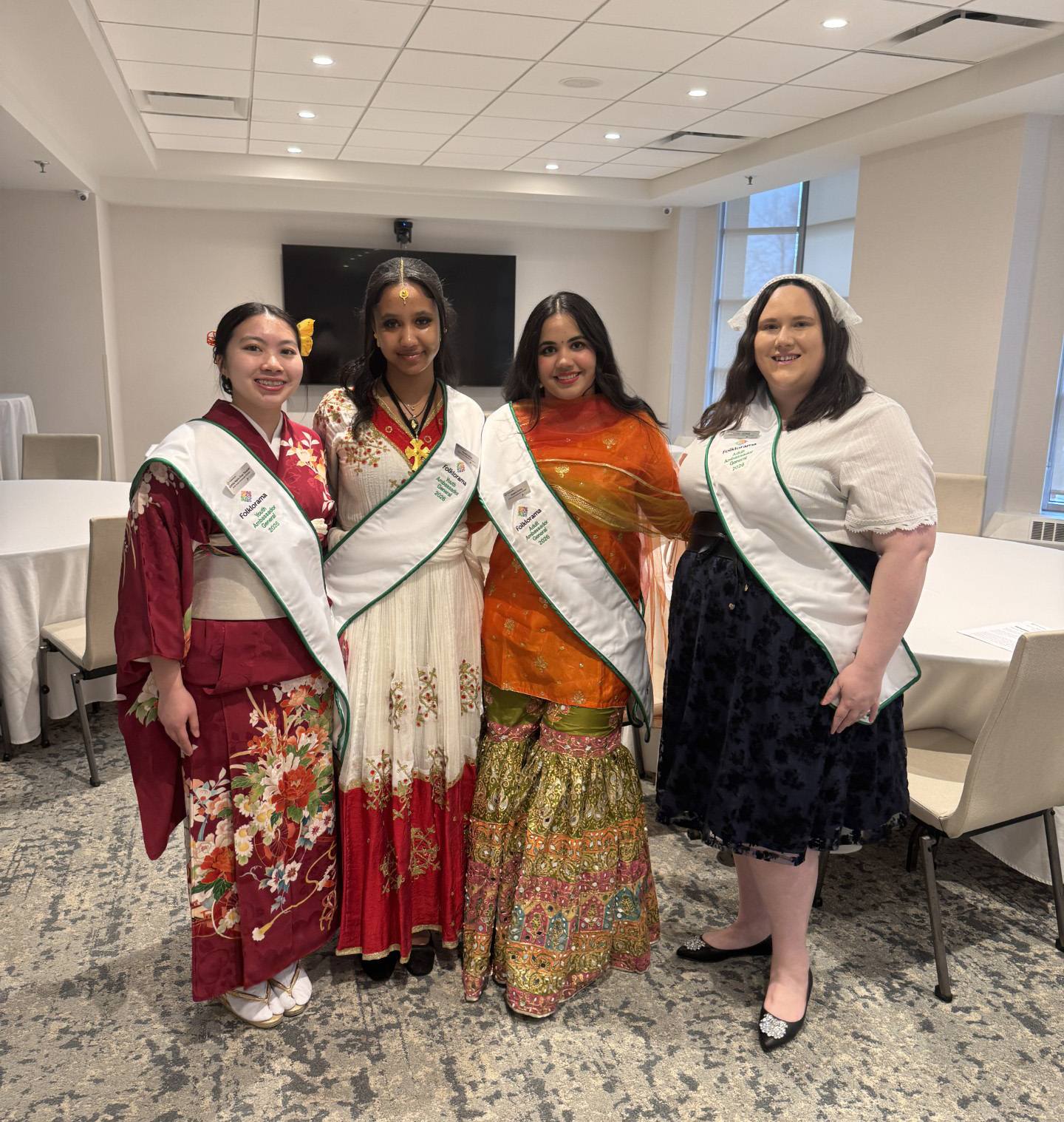 Four women in various cultural attire stand together in a line, smiling