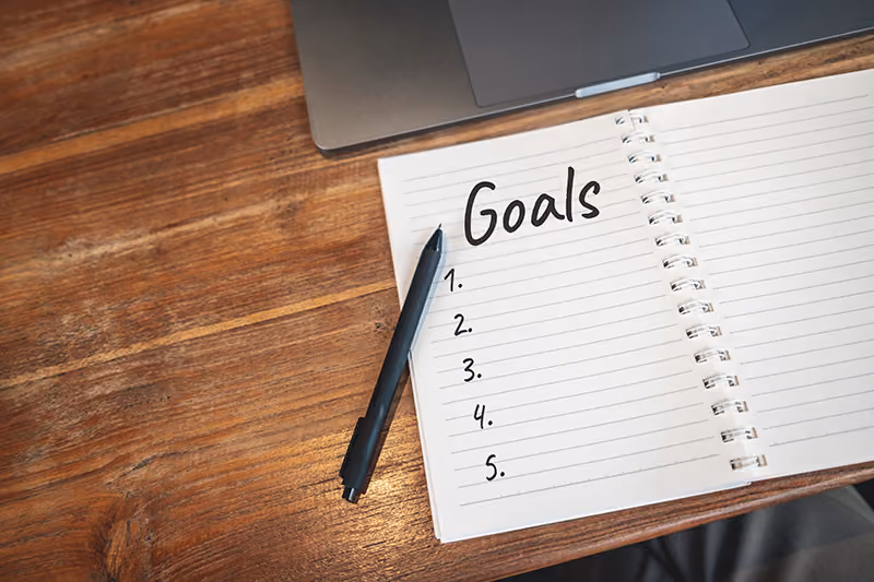 A lined spiral-bound notebook resting open with title "Goals" above a blank numbered list, sitting on a wooden table with a pen lying across the left page and a grey laptop partially in view