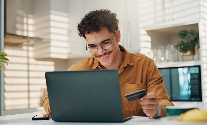 A man with glasses smiles down at a laptop screen, seated at a counter in a kitchen, a bank card in his left hand
