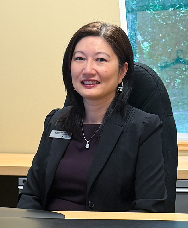 Woman with dark hair wearing a black blazer and necklace sitting at a desk in an office.