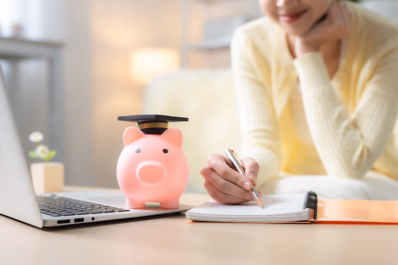 Someone in a pale yellow sweater writes in a notebook next to an open laptop and a pink piggy bank with a graduation cap