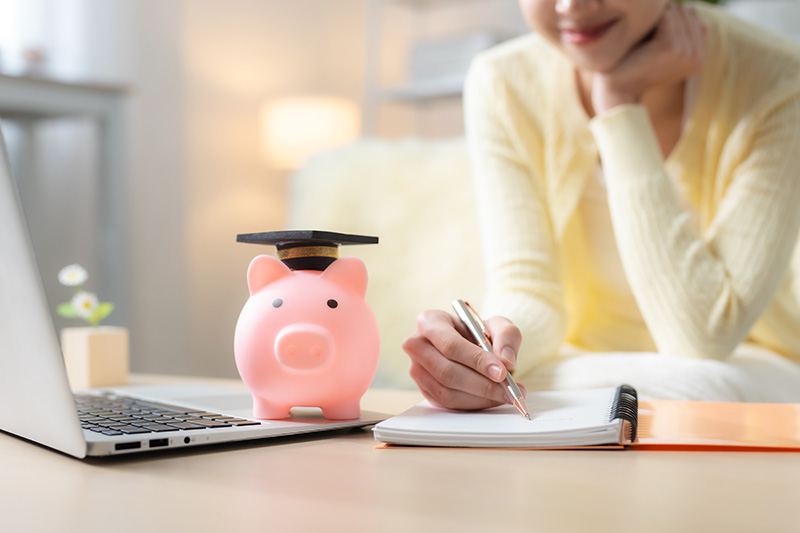 Someone in a pale yellow sweater writes in a notebook next to an open laptop and a pink piggy bank with a graduation cap