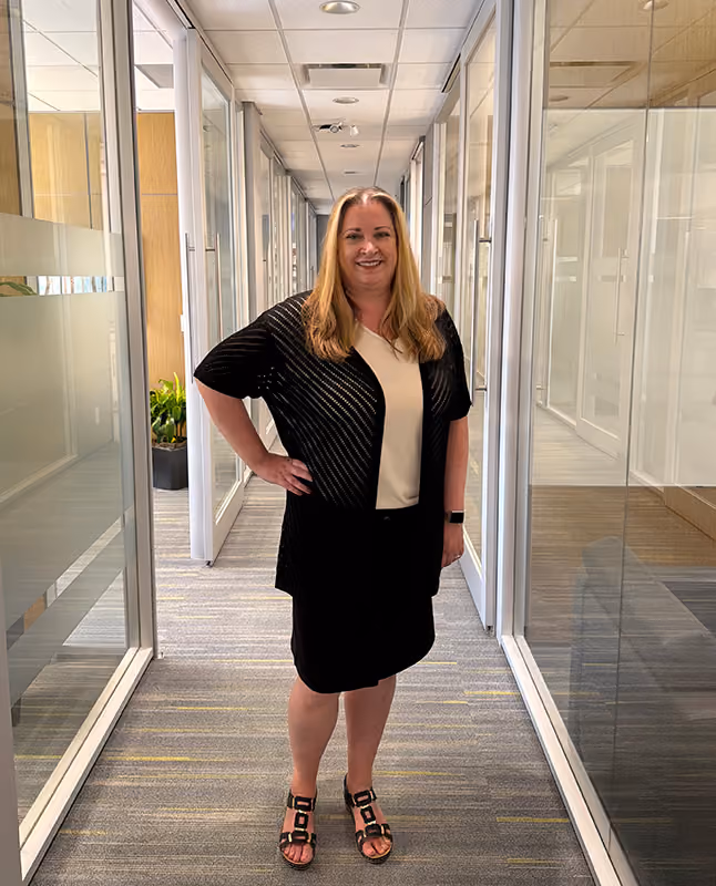 Smiling woman with long blonde hair wearing a beige top, black cardigan, black skirt, and black sandals standing in a bright, modern office hallway with glass walls and a gray carpet.