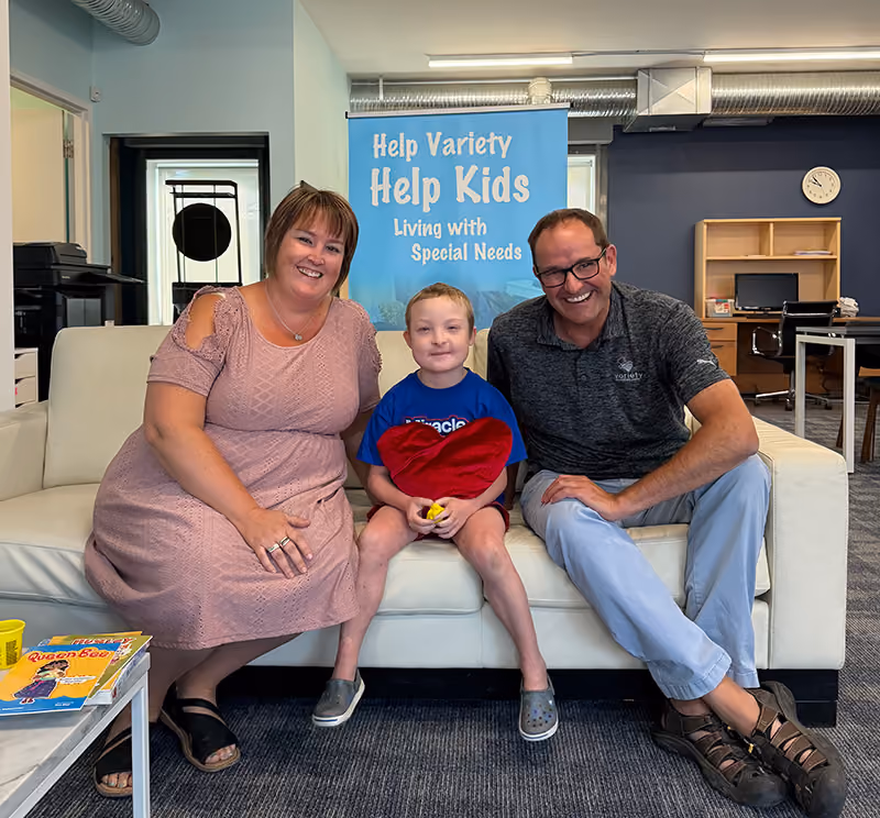 A woman, young boy, and man sit on a couch and smile in front of a sign that reads "Help Variety help kids living with special needs"