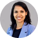 Professional headshot of a smiling woman with shoulder-length dark hair wearing a light blue blazer over a black top, posing against a plain light gray background in a circular frame.