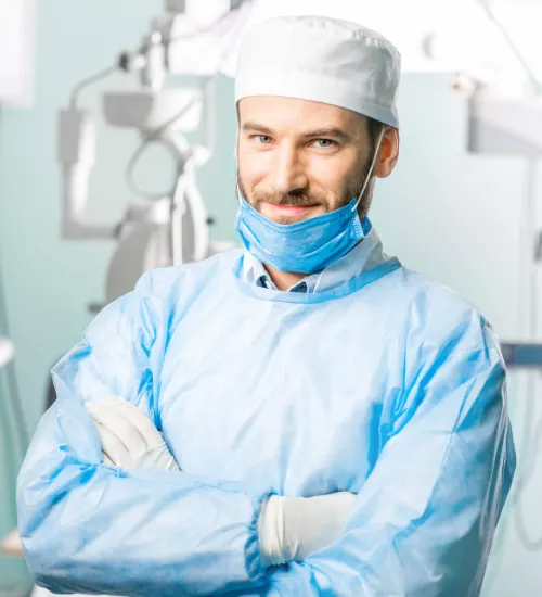 Male surgeon in blue scrubs, wearing a white cap and surgical mask below his chin, standing with arms crossed in a medical setting.