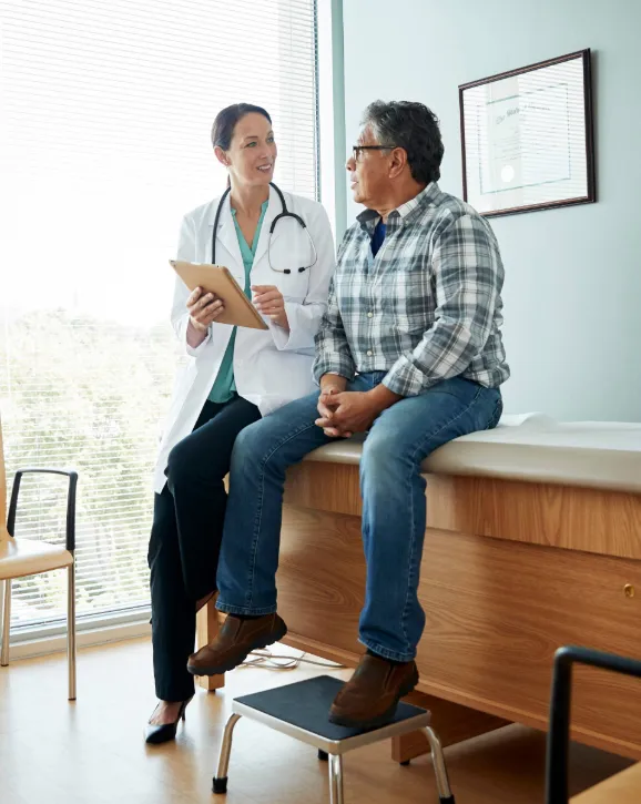 Female doctor in white coat holding a clipboard talking to an older male patient sitting on an exam table in a medical office.