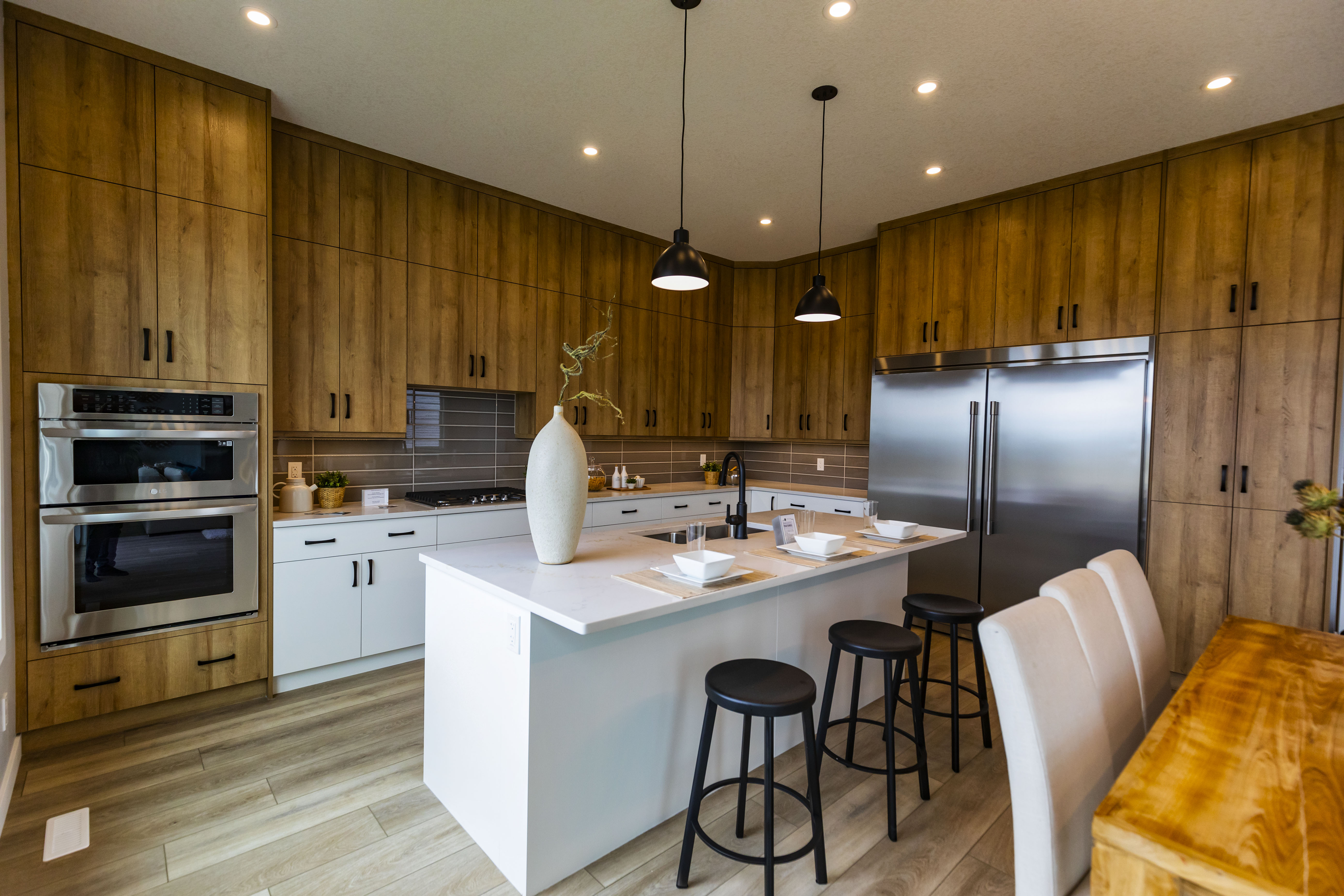 Modern kitchen with wooden cabinets, stainless steel double oven, large refrigerator, white island with three black stools, and pendant lights above.