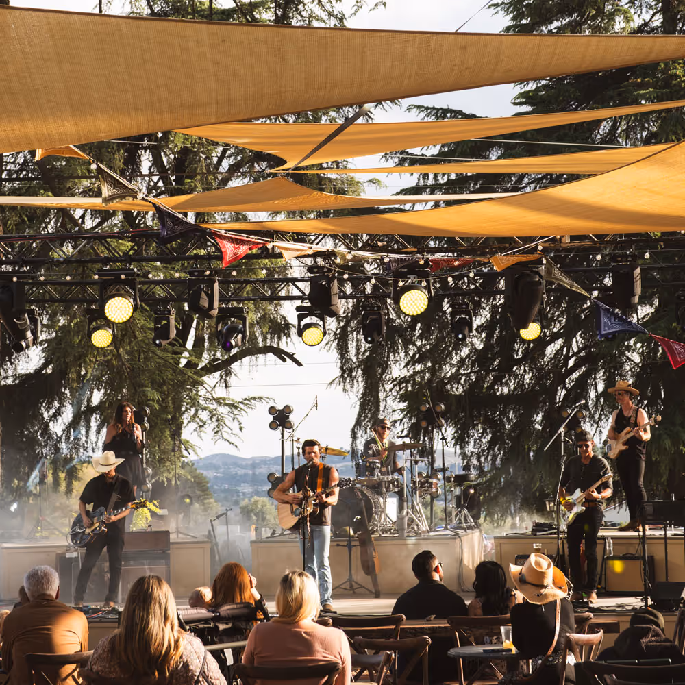 A country music band performs on a rustic wooden stage marked "123 FARM" as people dance at sunset.