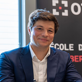 Smiling man with brown hair wearing a blue suit and white shirt in front of a black banner with white and red text.