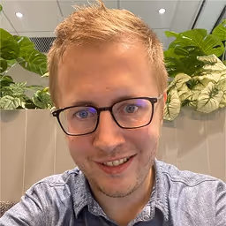 Smiling young man with glasses and short blond hair wearing a blue shirt, sitting indoors with green plants in the background.