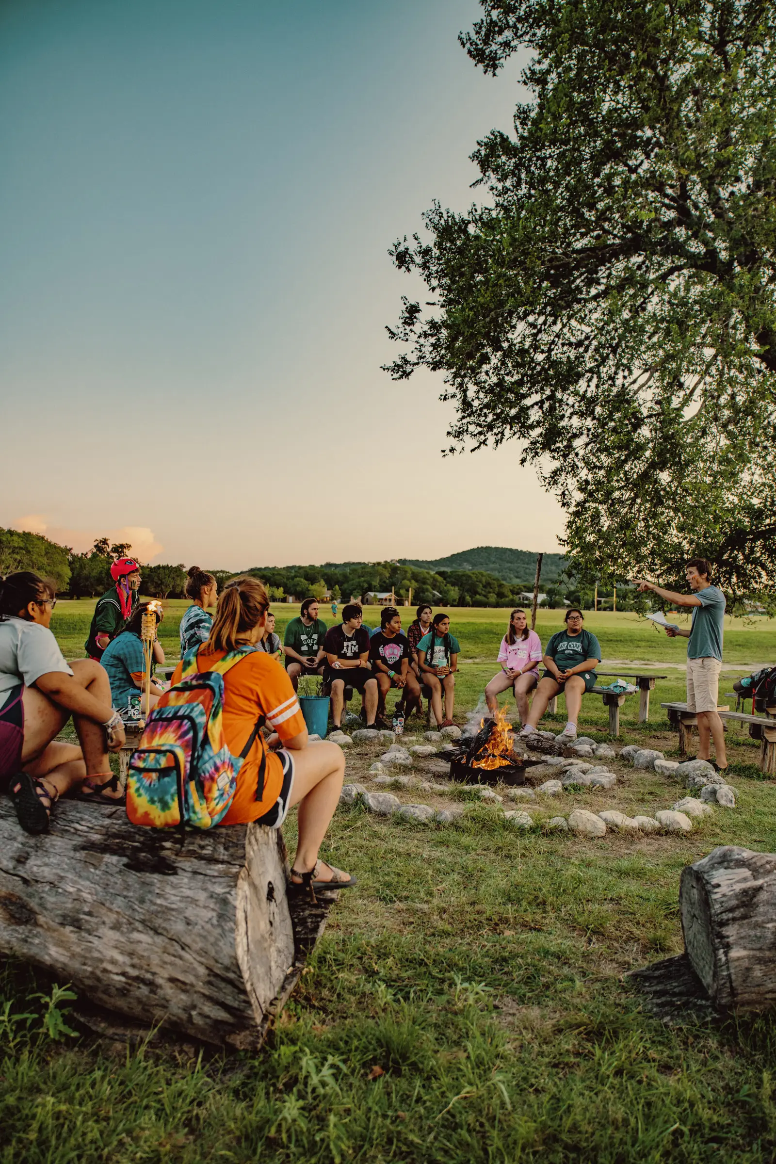 Group of people sitting around a campfire ring in a grassy field, listening to a speaker near a tree.