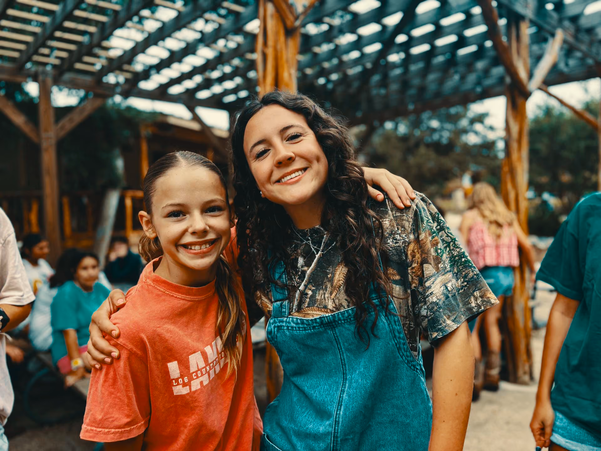 Two smiling girls stand close together under a wooden pergola; one girl with long dark curly hair wears a camouflage shirt and denim overalls, the other with long straight blonde hair wears a red t-shirt, with blurred people and greenery in the background.