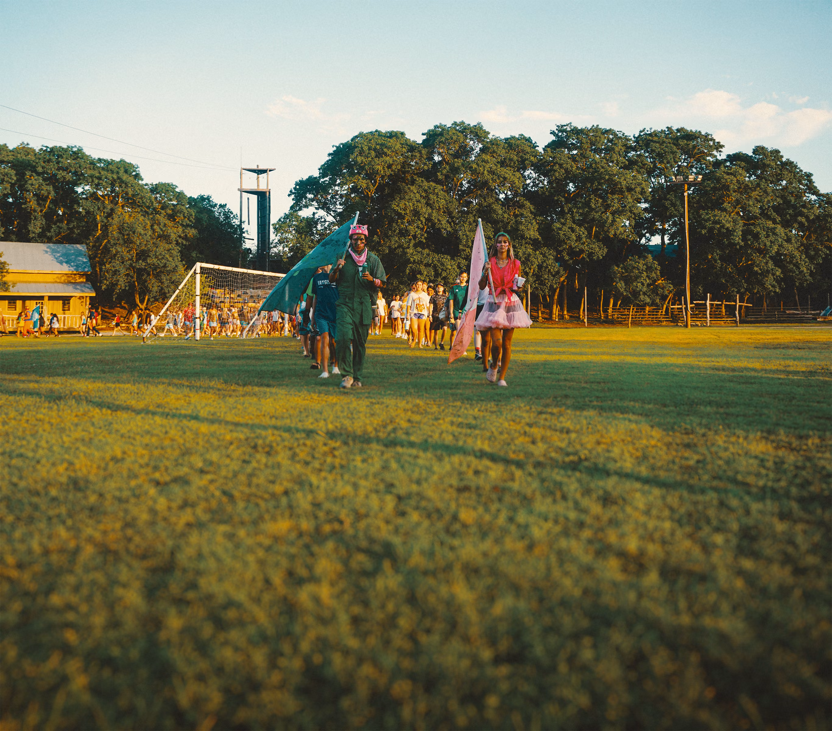 Group of people walking on grassy field with two individuals at the front holding flags, one dressed in a green outfit with a pink hat and the other in a pink dress, with trees and a soccer goal in the background under a blue sky.