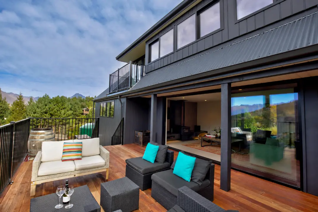 Broad sliding doors connect the living room to a furnished timber deck beneath the upper storey of the dark clad house.