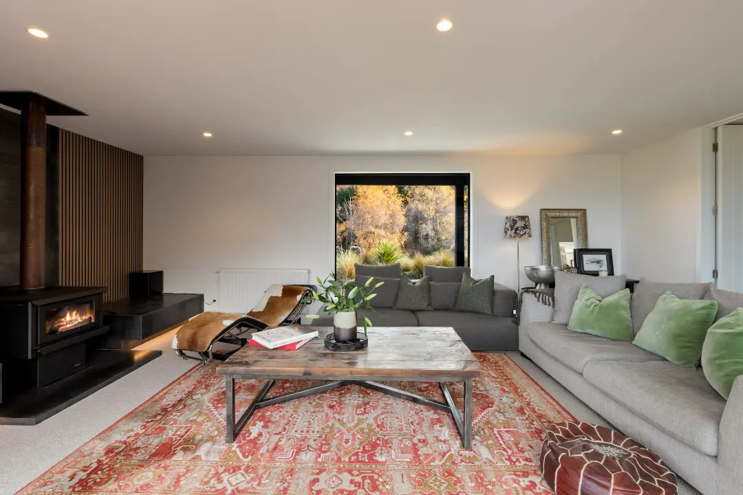 Living room with grey sectional seating, timber lounge chairs, a patterned rug and a wood burner against a timber feature wall.