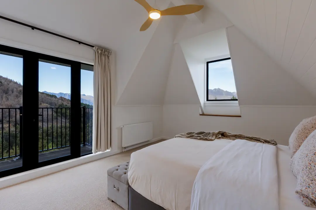 Bedroom beneath a pitched ceiling, with a dormer window and glazed balcony doors overlooking the mountains.