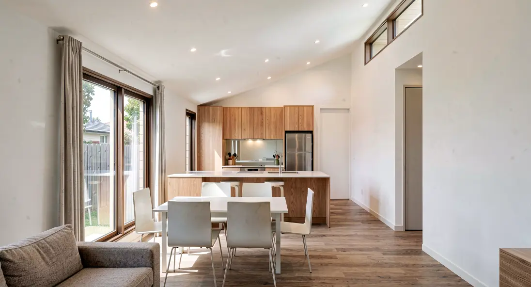 Open plan kitchen and dining area in Modal Architecture's Takahē Three home, featuring natural timber cabinetry, a breakfast bar and a sloping ceiling.