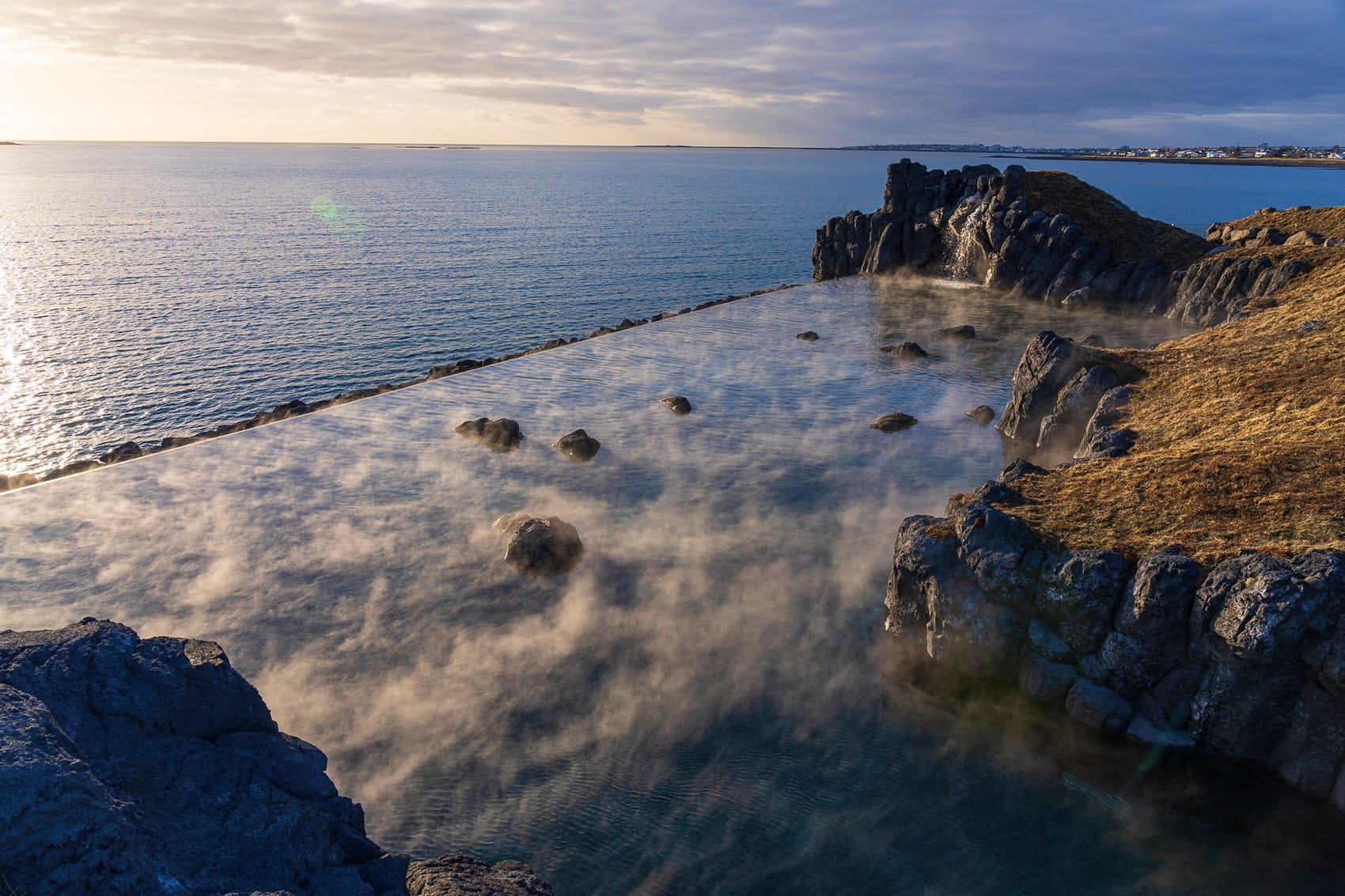 Rocky shoreline with steam rising from geothermal water along Iceland's iconic Golden Circle, meeting the calm ocean under a partly cloudy sky at sunset or sunrise—an unforgettable tour moment.