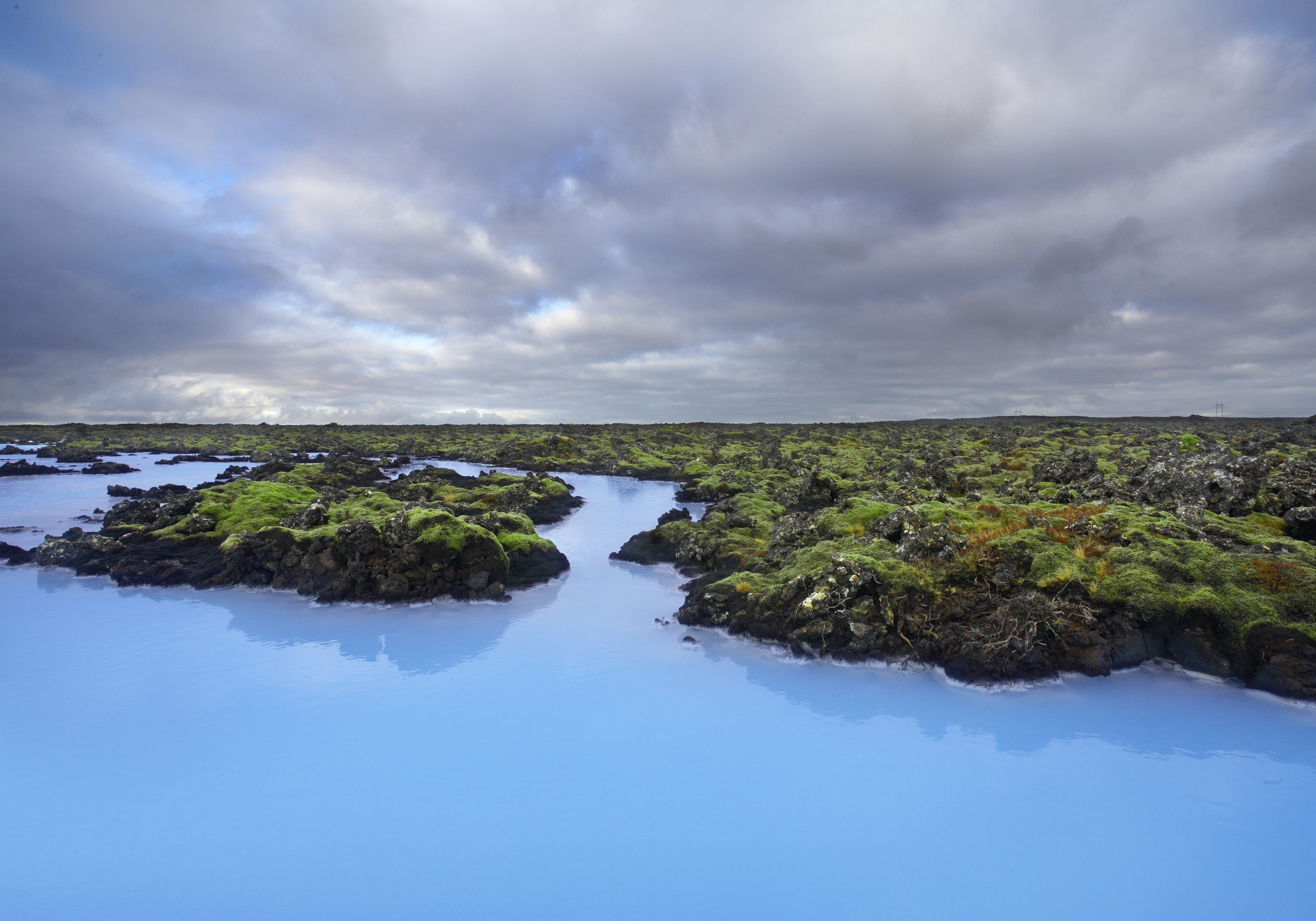 Blue geothermal pools surrounded by dark rocks and patches of green moss under a dramatic, cloudy sky in Iceland’s wide, open landscape—a stunning sight along the Golden Circle tour.