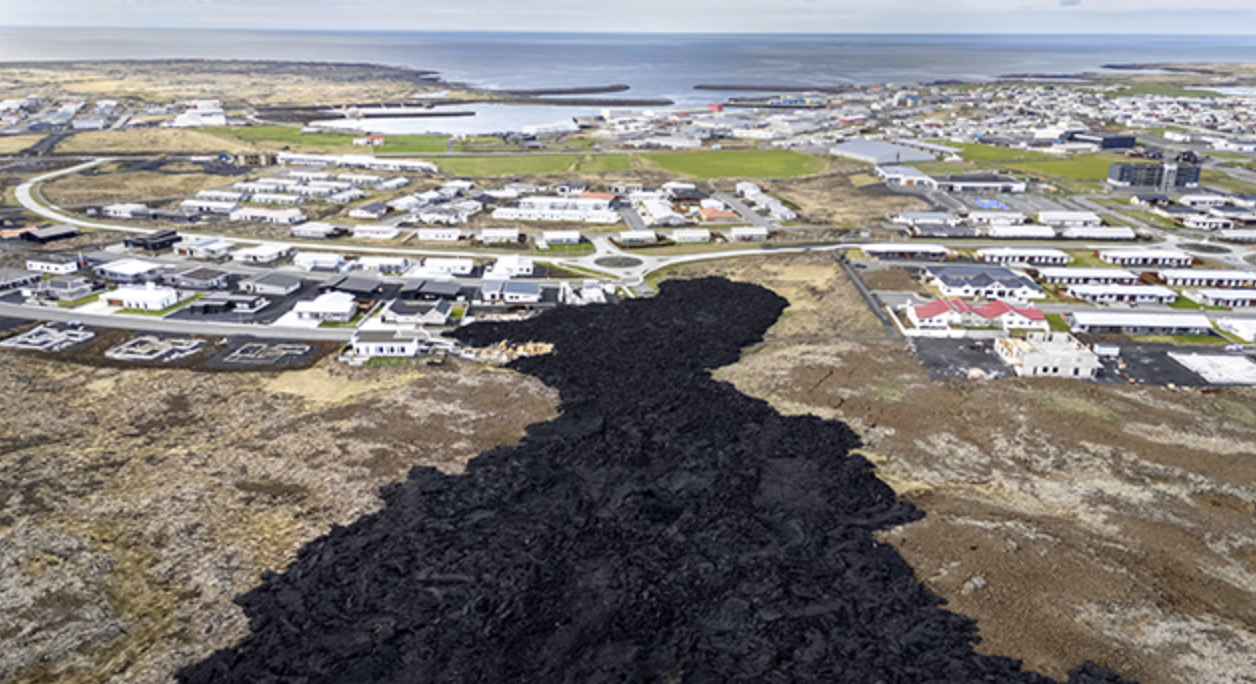 Aerial view of a town along Iceland’s coast, where dark lava flow cuts through grassy land near homes—an extraordinary sight often witnessed on a Golden Circle tour under a partly cloudy sky.