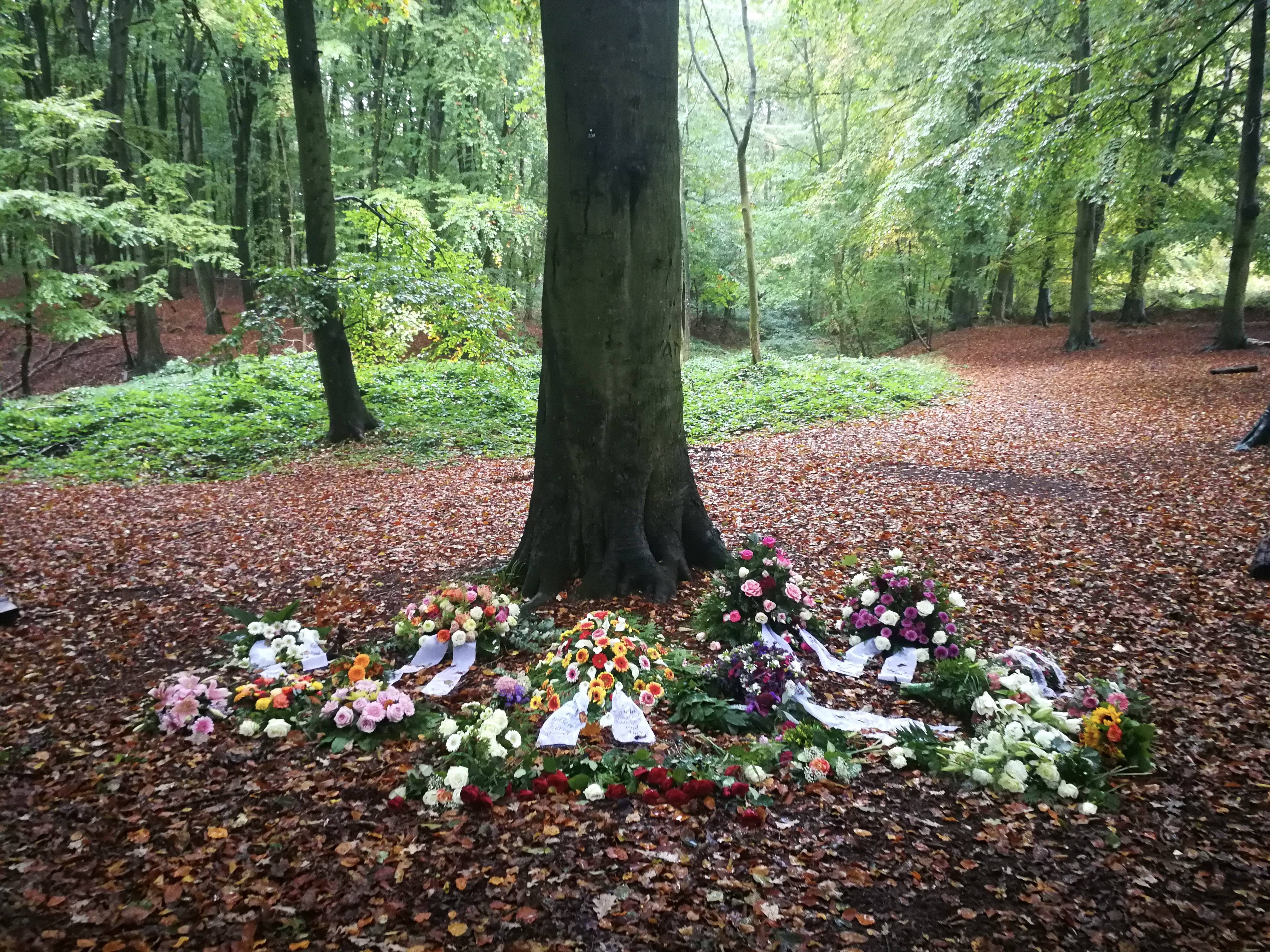 Showing a grave in the woods, decorated with flowers. Showing all green trees in the background.