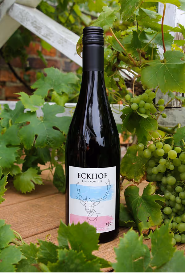 Eine Weinflasche mit der Aufschrift ‚Eckhof Einer von hier‘ steht auf einem Holztisch, umgeben von grünen Weinblättern und Trauben.