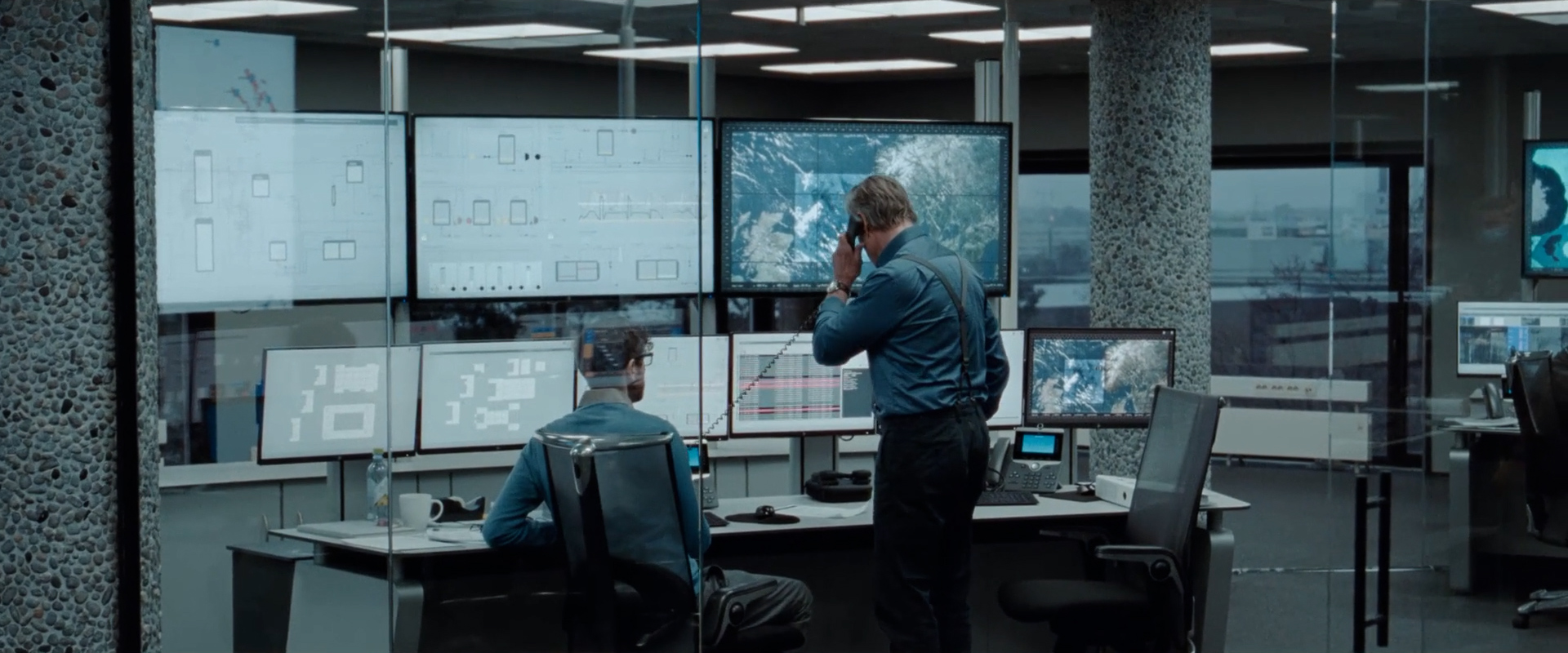 Two men in an office surrounded by multiple monitors displaying maps, data, and graphs, one is seated and the other is on the phone.