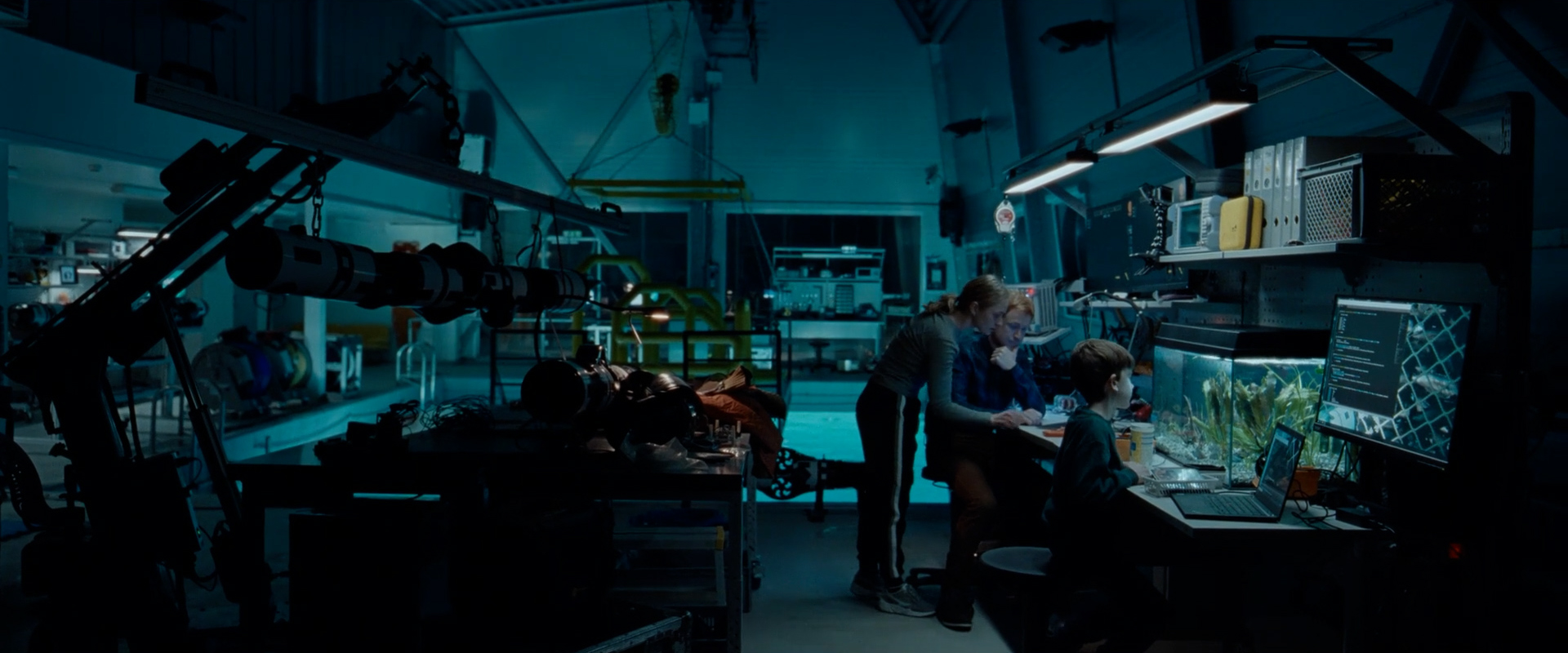 Three people in a dimly lit laboratory or workshop, working at a desk with a laptop, large monitor, and an aquarium filled with plants.