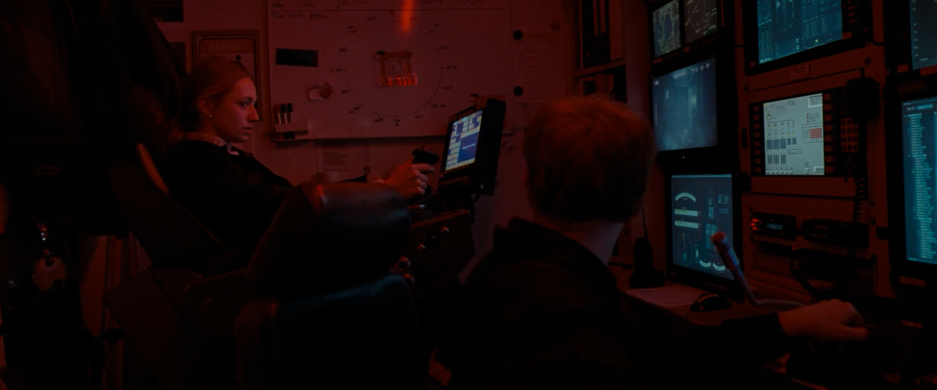 Two people operating controls and monitors in a dimly lit control room with red lighting.