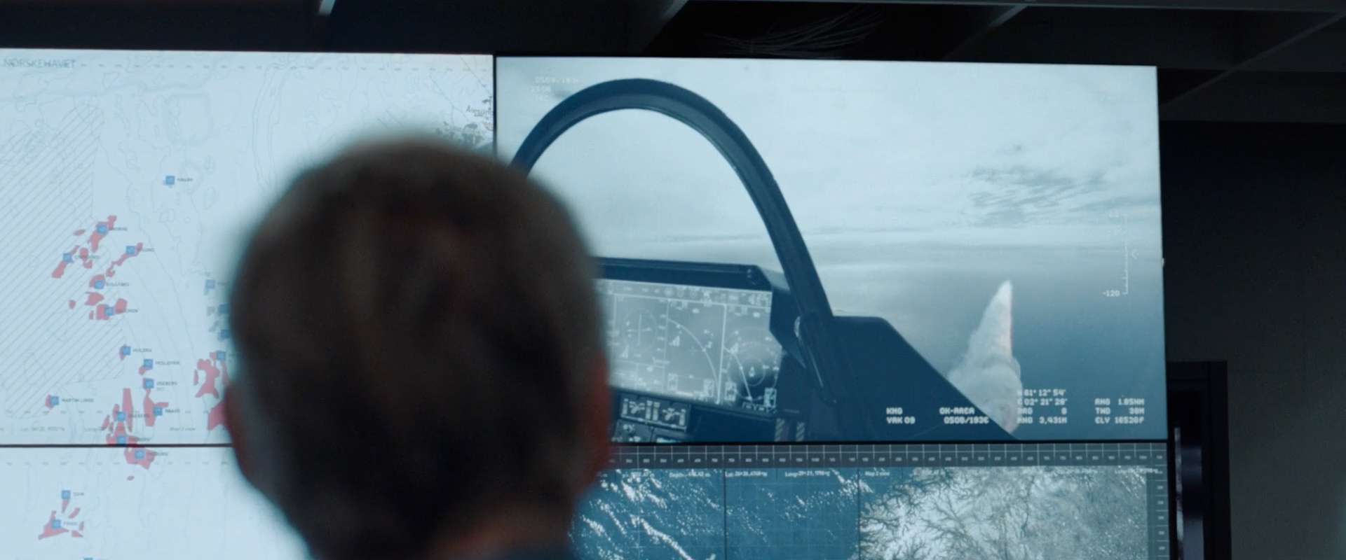 Back of a person's head viewing large screens displaying a map and a cockpit view of a fighter jet flying over an ocean.