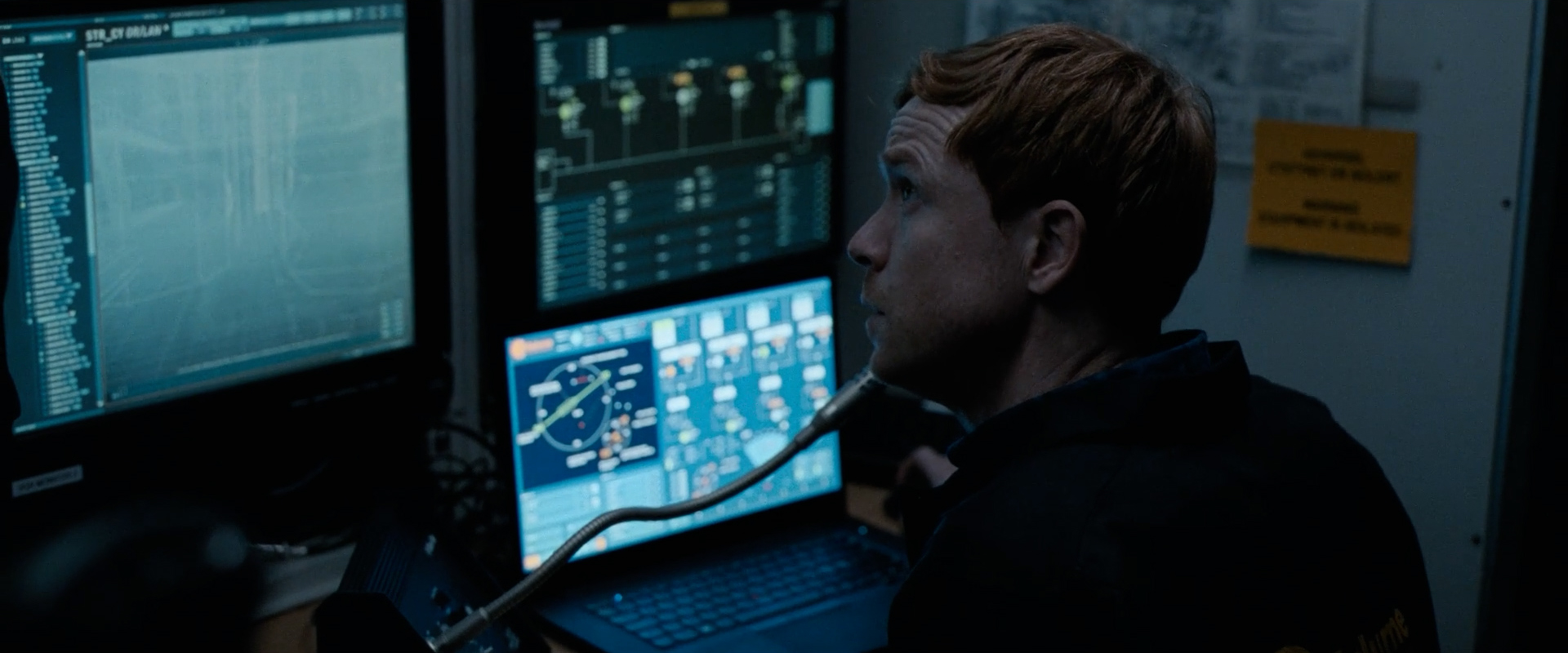 Man with reddish hair using a microphone and looking at multiple computer screens displaying technical data.
