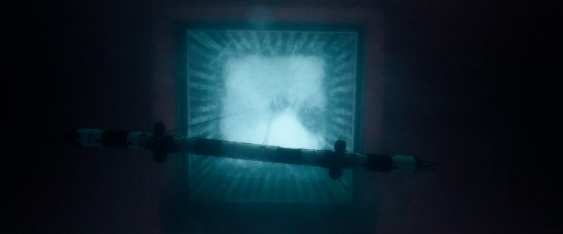 Underwater view of a horizontal cylindrical device suspended by cables above a lit square structure.