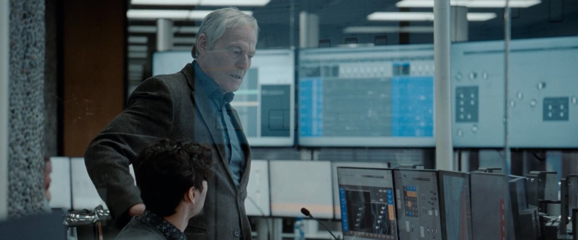 Older man standing and talking to a younger man seated in front of computer monitors in a control room.
