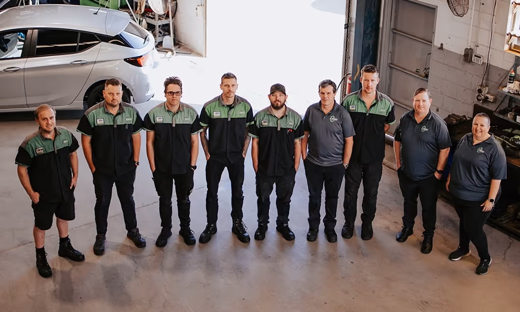 Group of nine people in uniform standing in a warehouse garage next to a silver car.