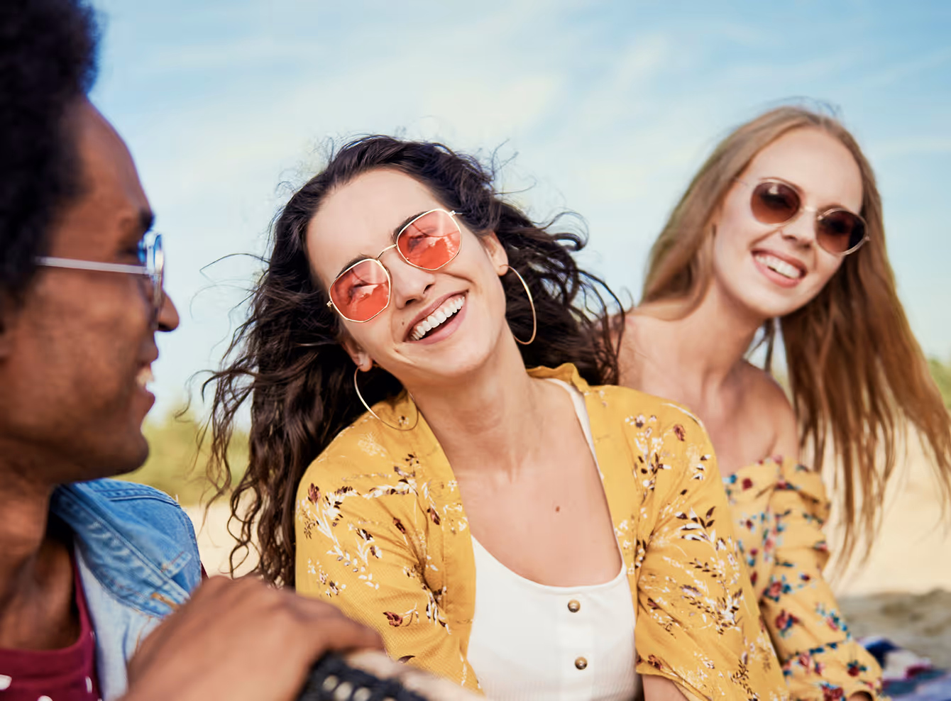Three young adults wearing sunglasses smiling and enjoying themselves outdoors with a clear blue sky in the background.