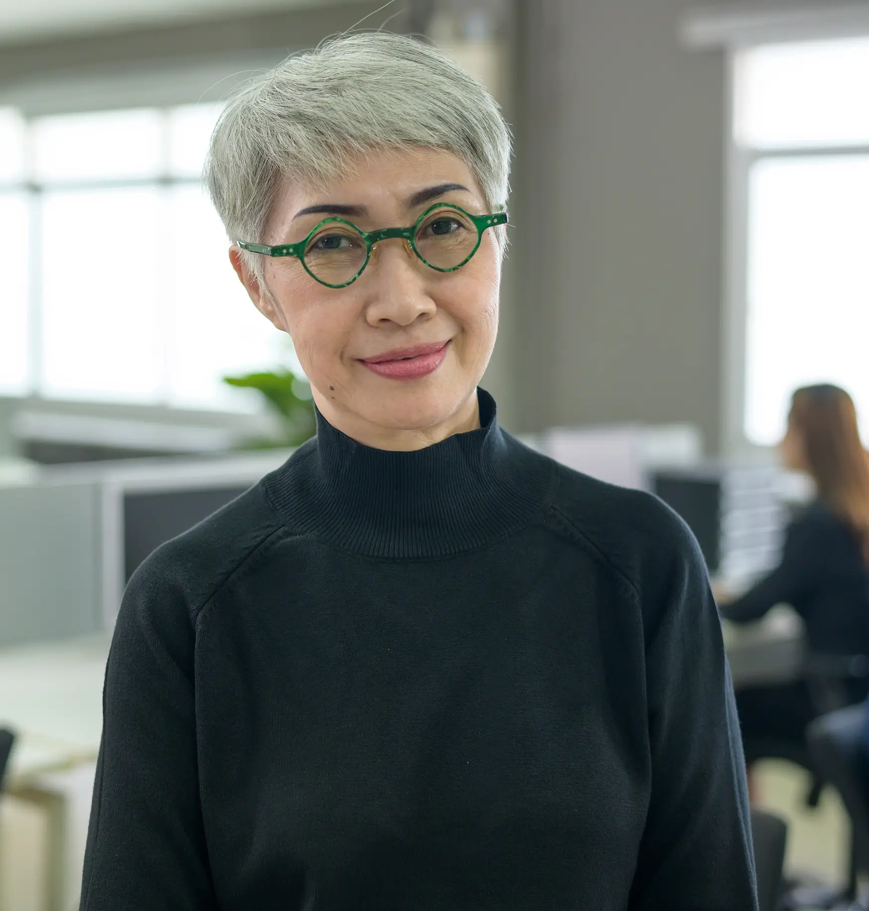 Mujer mayor sonriente con cabello corto gris y gafas redondas verdes, vestida con un jersey de cuello alto negro en una oficina luminosa.