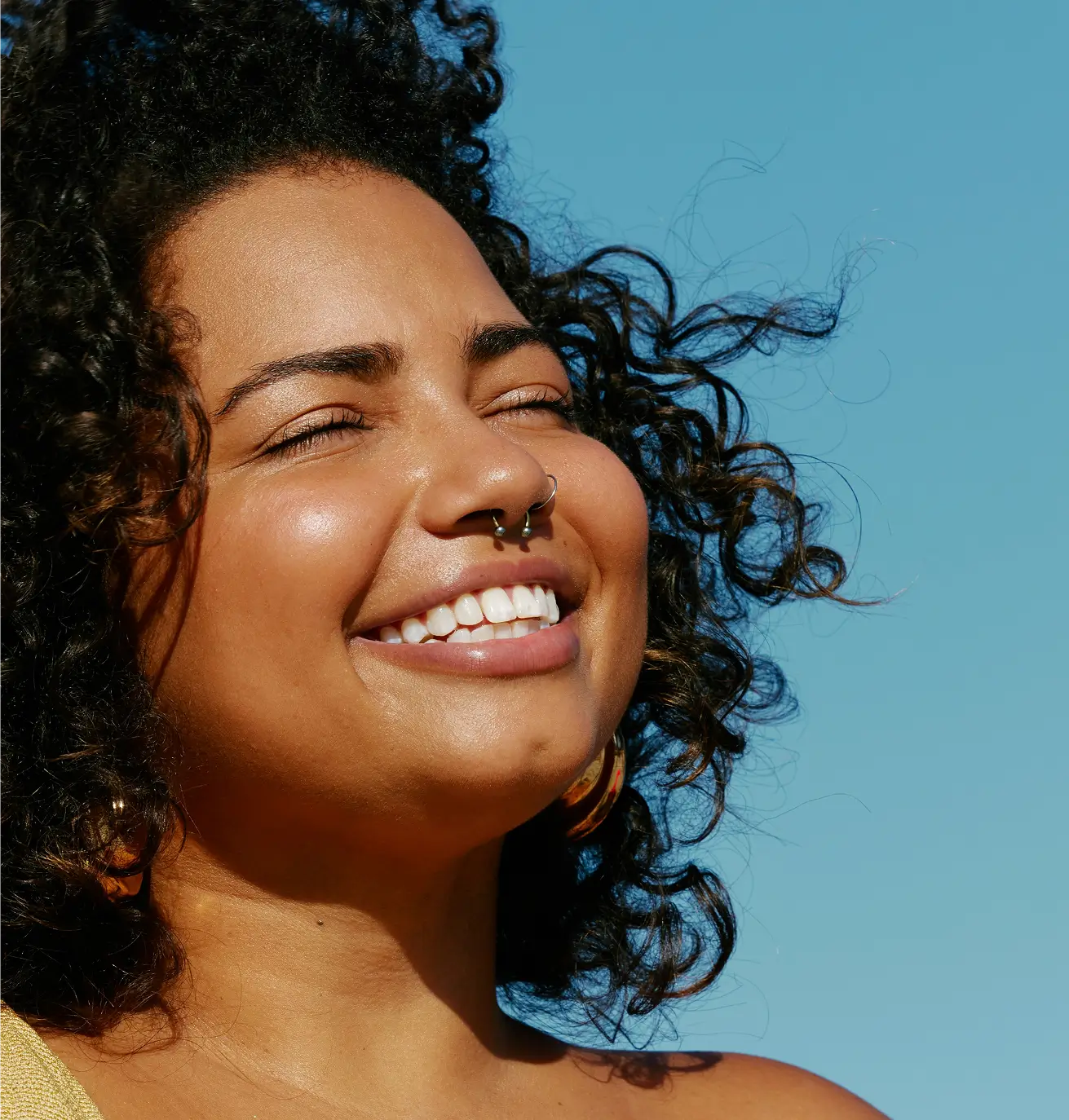 Primer plano de una mujer sonriente con cabello rizado y un piercing en la nariz contra un cielo azul claro.