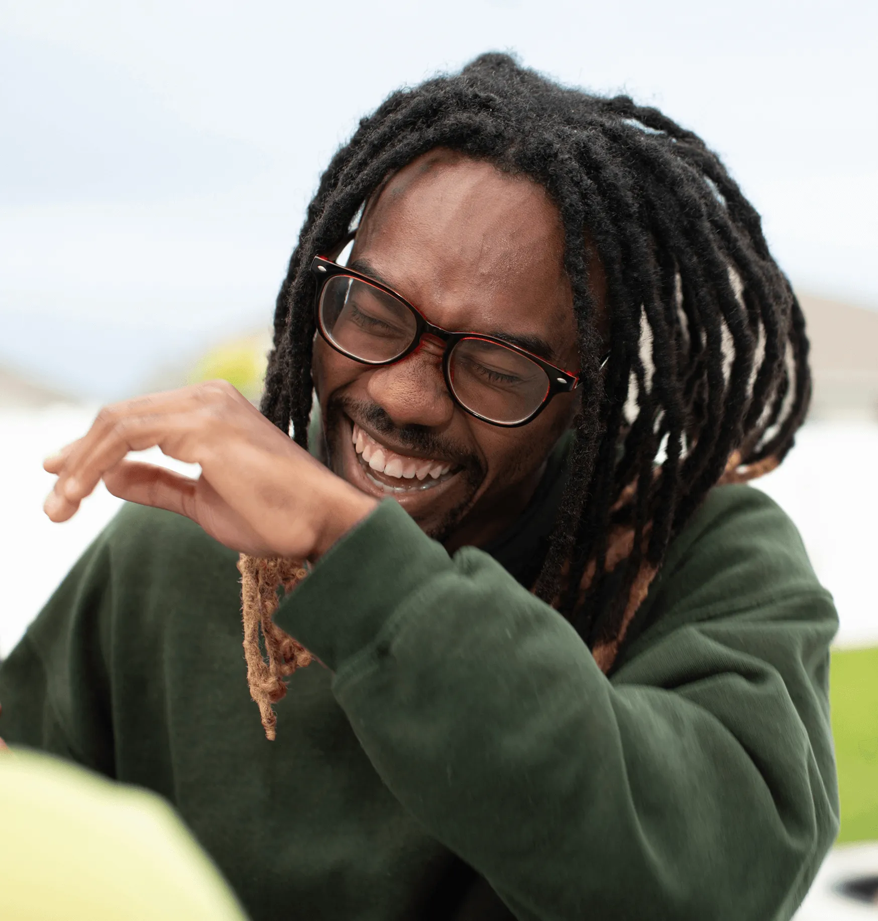 Hombre sonriente con rastas y gafas, vestido con una sudadera verde, cubriéndose la boca con el brazo mientras se ríe.