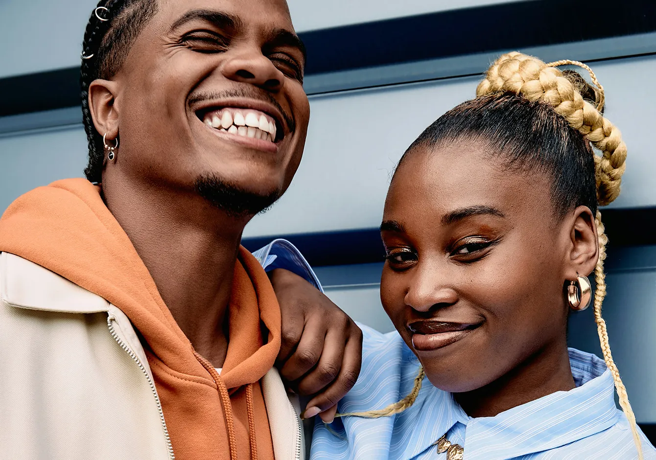 Primer plano de un hombre sonriente con una sudadera naranja con capucha y una mujer con trenzas y una camiseta azul, posando juntos.