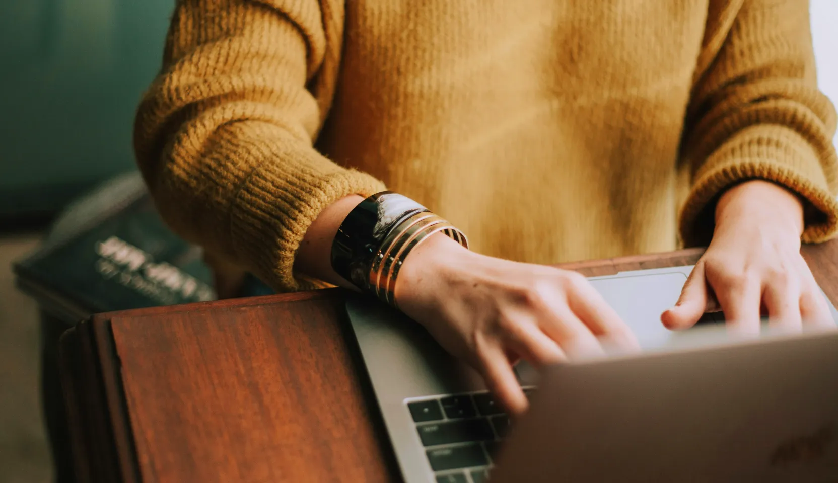 Persona con un jersey marrón escribiendo en un ordenador portátil con una gruesa pulsera plateada y negra en la muñeca izquierda.