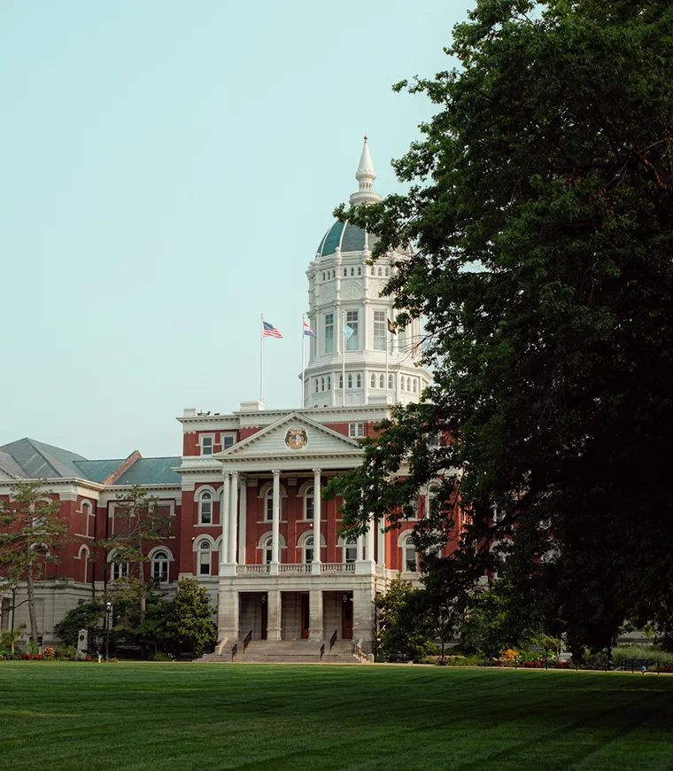 Edificio gubernamental de ladrillo rojo con columnas blancas y una torre abovedada, parcialmente enmarcado por árboles, con banderas ondeando en el tejado.