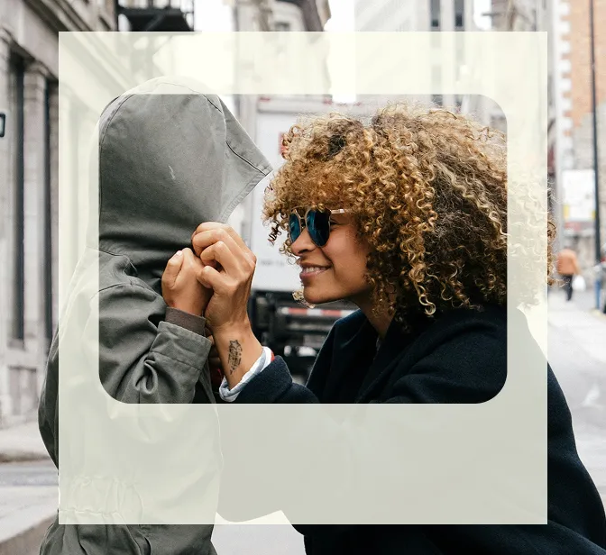 Una mujer con el pelo rizado y gafas de sol que, con aire juguetón, le arregla la capucha de la chaqueta a un niño en una calle de la ciudad.