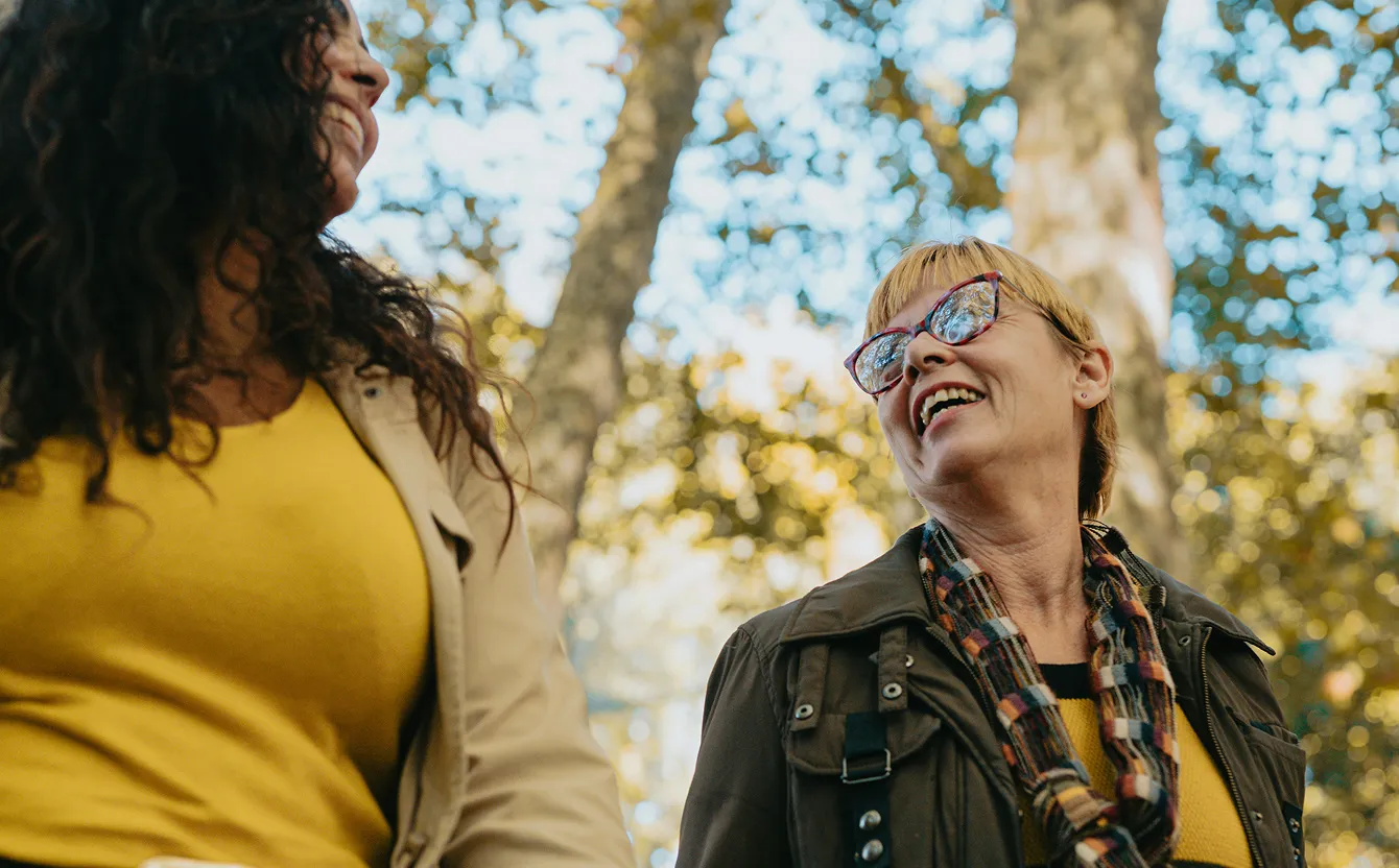 Two women smiling and talking outdoors with trees and sunlight in the background.